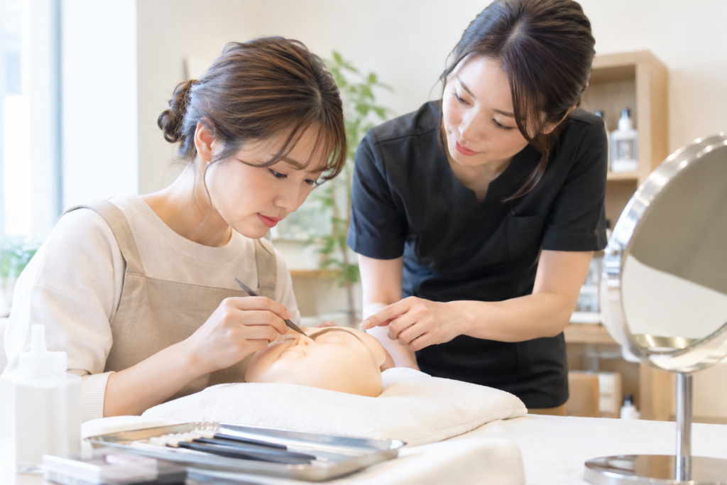 エステサロンのような室内で、2人の女性が施術用ベッドの上で顔まわりの練習をしている