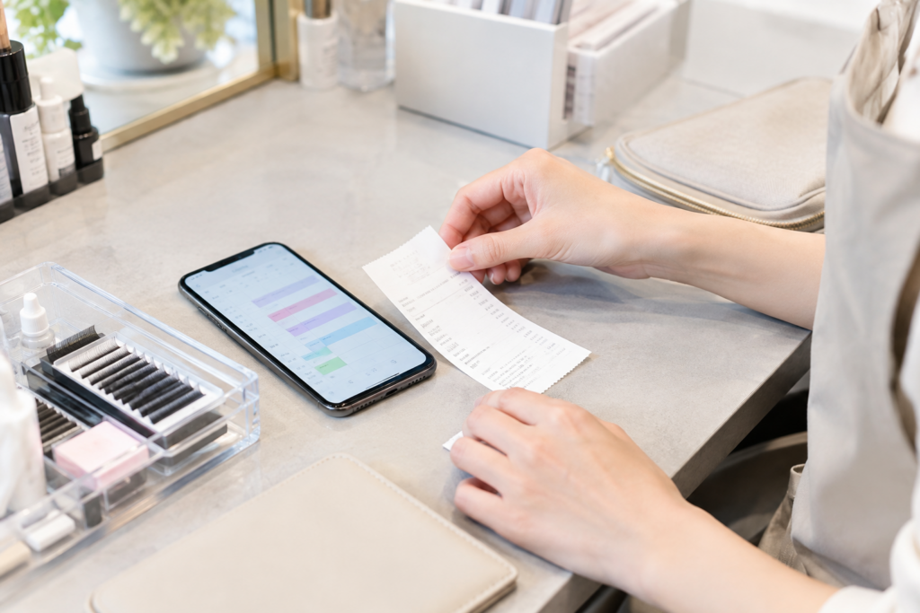 サロンの机でスマートフォンと紙の明細を見比べる手元