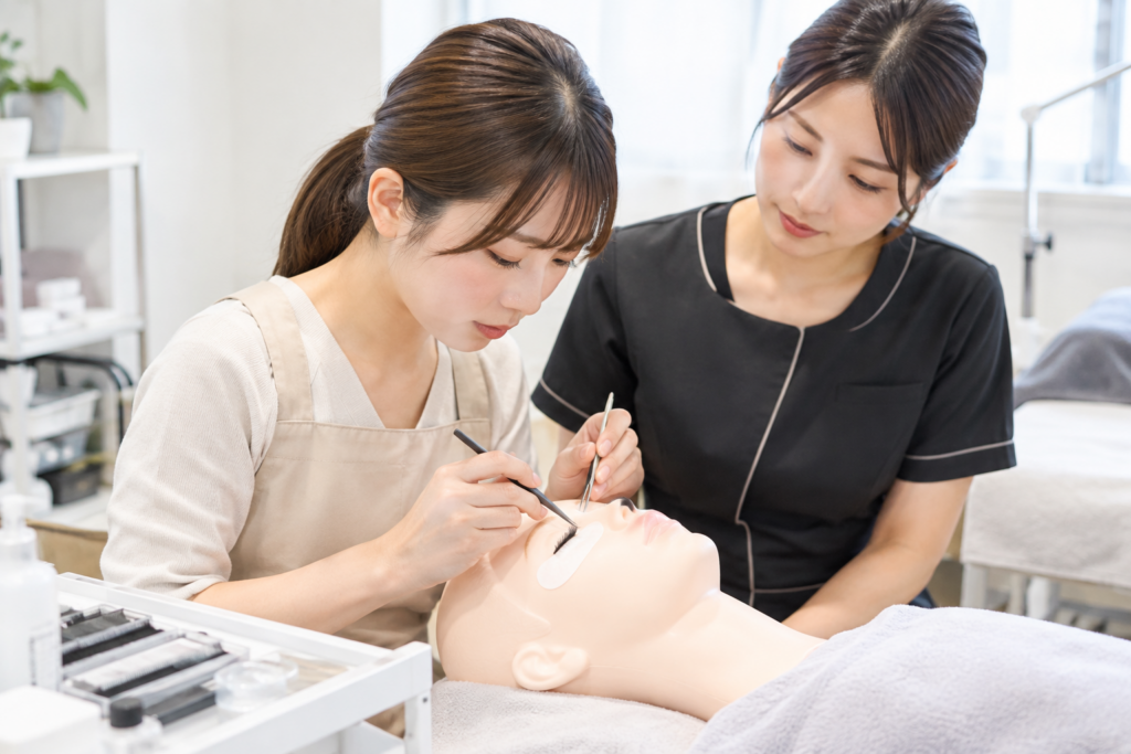 施術用マネキンの目元で、2人の女性がまつげ施術の練習をしている