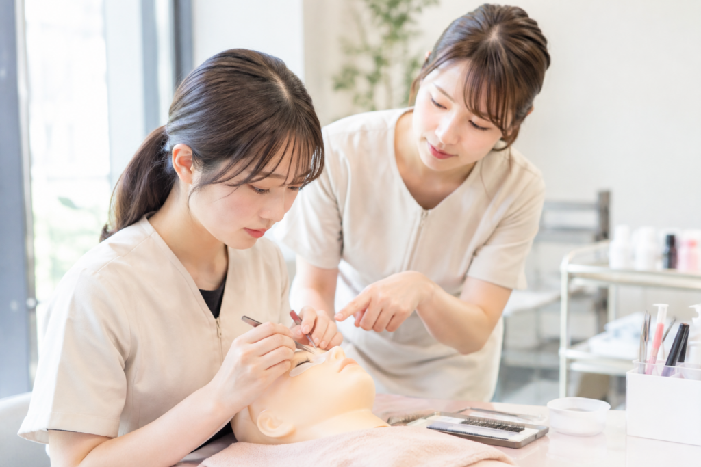 エステサロンで、女性スタッフが横たわる女性にまつ毛の施術を教えている