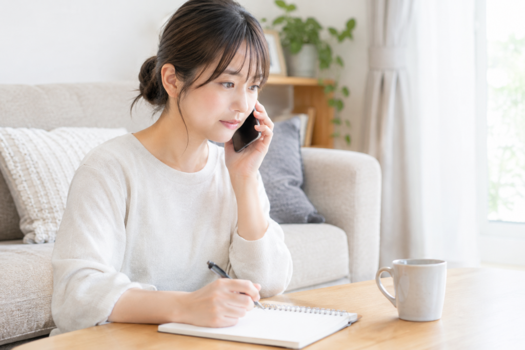 女性が自宅でスマートフォンで通話しながら、ノートにメモを取っている